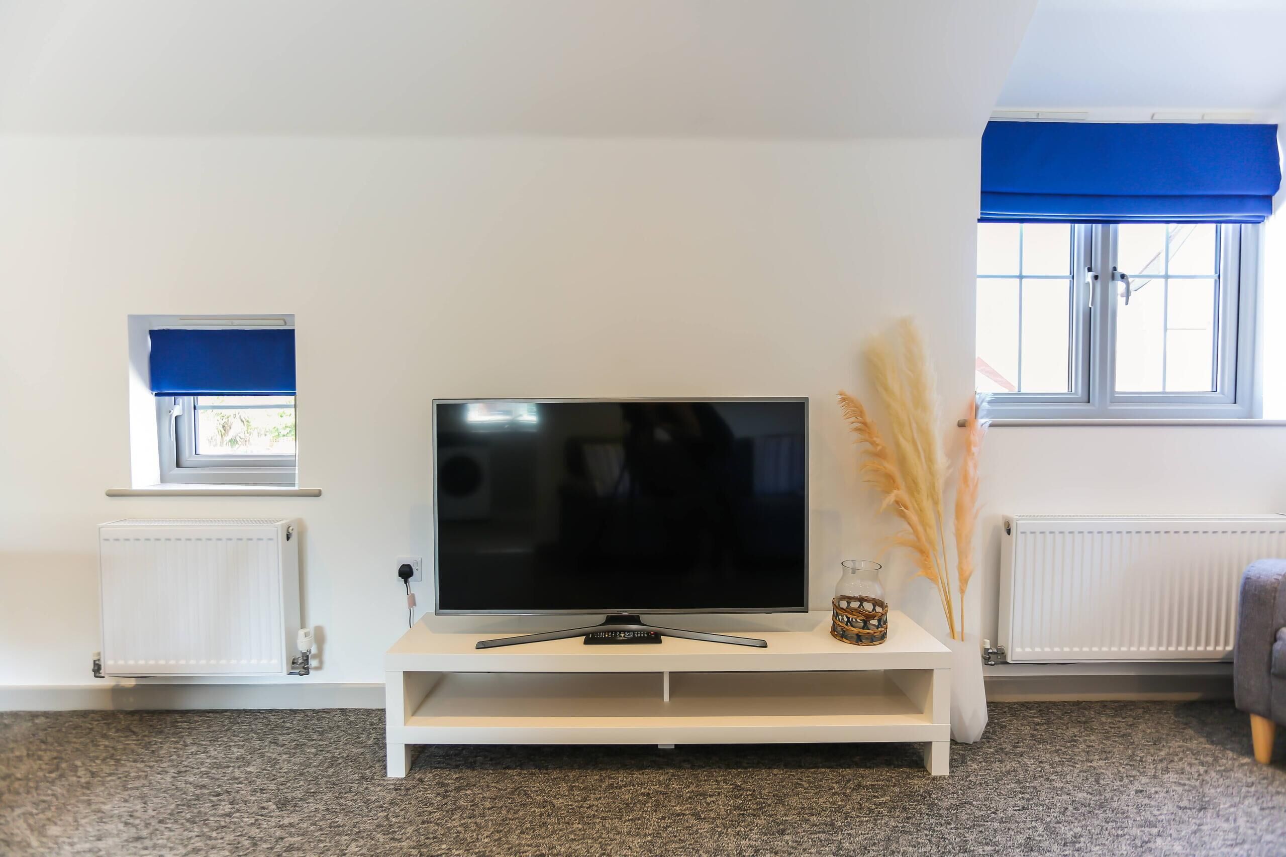 Una sala de estar con un televisor de pantalla plana sobre un soporte blanco, una mesita con un frasco de vidrio y una ventana con persianas azules.