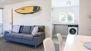 A modern living room with a grey sofa, a surfboard on the wall, a washing machine, and a window with blinds. - Shorebreak - Bracklesham Bay Apartment (Chichester)