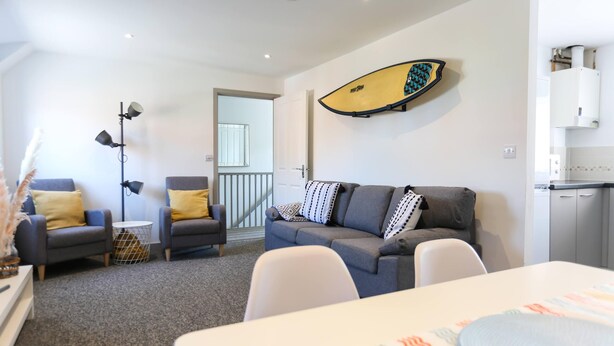 A modern living room with a grey sofa, two armchairs, a dining table, and a surfboard mounted on the wall. - Shorebreak - Bracklesham Bay Apartment (Chichester)