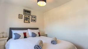 A neatly made bed with towels, a bedside table, and a wall with framed pictures. - Shorebreak - Bracklesham Bay Apartment (Chichester)