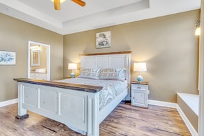 A bedroom with a wooden bed frame, two bedside tables, a ceiling fan, and a painting on the wall.