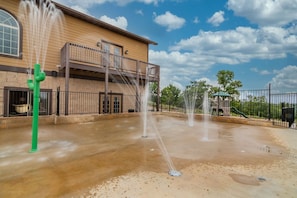 A water feature with multiple fountains in a courtyard, a building with a balcony, and a playground with slides in the background.