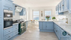 A modern kitchen with light blue cabinets, a central island, and a dining area with white chairs. - Comfort Hill - Luxury, hot tub, sea views (Weymouth)
