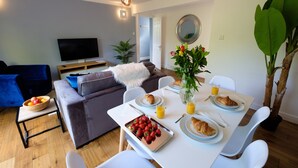 En moderne stue med en grå sofa, et fjernsyn og et spisebord dækket med morgenmad.