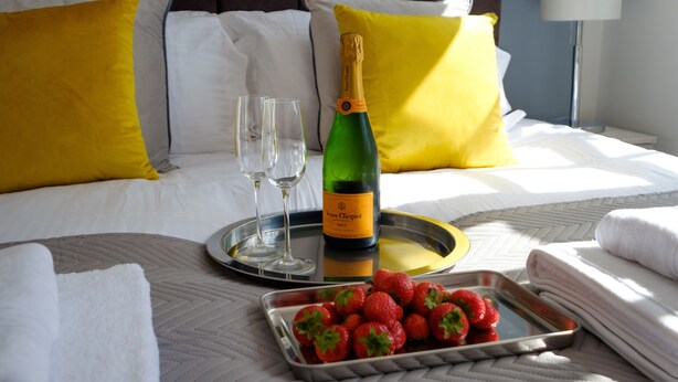 En pænt redt seng med hvide og gule puder, en flaske champagne, to glas og et fad med jordbær.