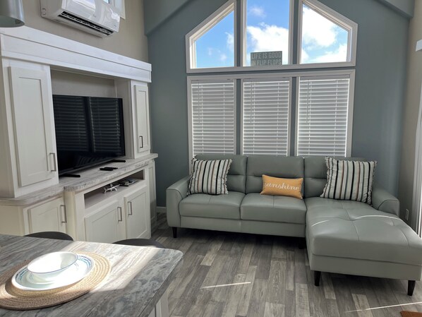 A modern living room with a grey sectional sofa, a flat-screen TV, and a large window with blinds.