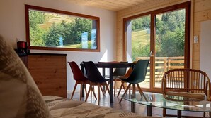 Een gezellige kamer met een bed, een eettafel met stoelen en grote ramen met uitzicht op groen.