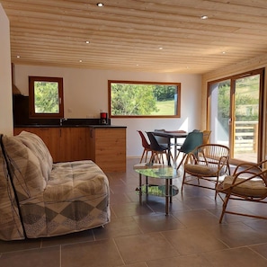 Uma sala de estar moderna com um sofá, uma mesa de centro de vidro e cadeiras de vime. Há uma cozinha com uma pia e uma janela com vista para um jardim.