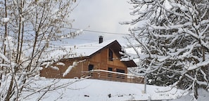 Uma cabana de madeira com telhado inclinado, rodeada por árvores cobertas de neve e um teleférico ao fundo.
