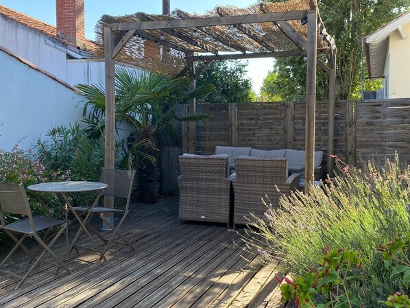 A wooden deck with wicker furniture and a table, surrounded by plants and a wooden fence.