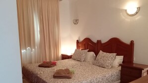 A neatly made bed with a patterned bedspread, two pillows, and a folded towel. There is a bedside table with a lamp and a nightstand with a lamp.