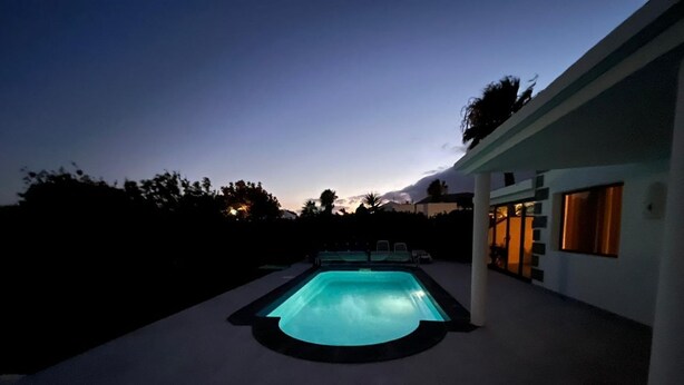 A swimming pool with a light blue water feature, surrounded by a concrete deck and a modern house with large windows.