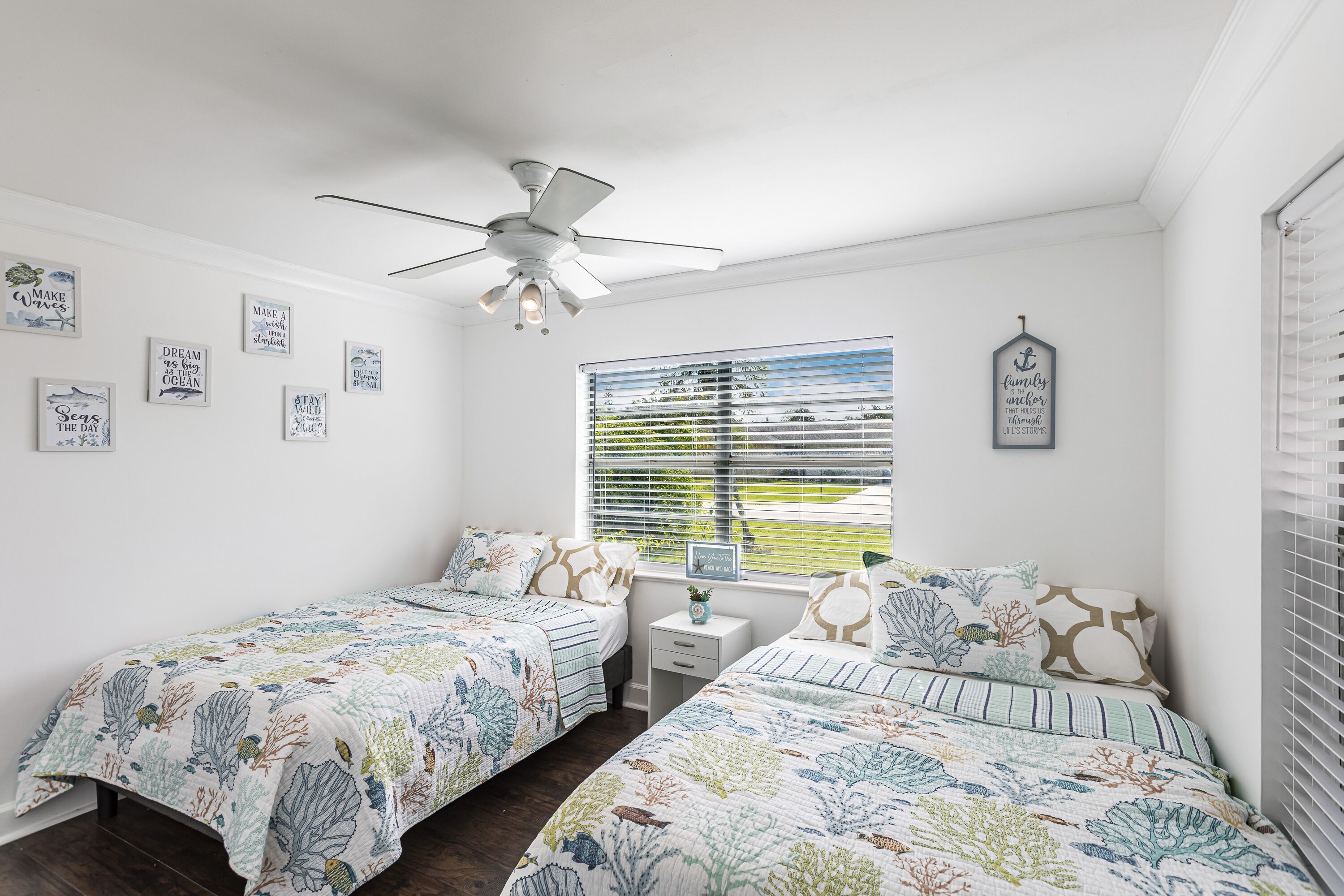 A bedroom with two single beds, a ceiling fan, a window with blinds, and several wall art pieces.