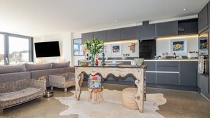 Un salon moderne avec un canapé gris, des fauteuils en osier, une table basse avec des magazines et une grande fenêtre.