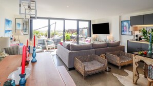 Un salon moderne avec un grand canapé, des fauteuils en osier, une table à manger en bois et un téléviseur à écran plat fixé au mur.