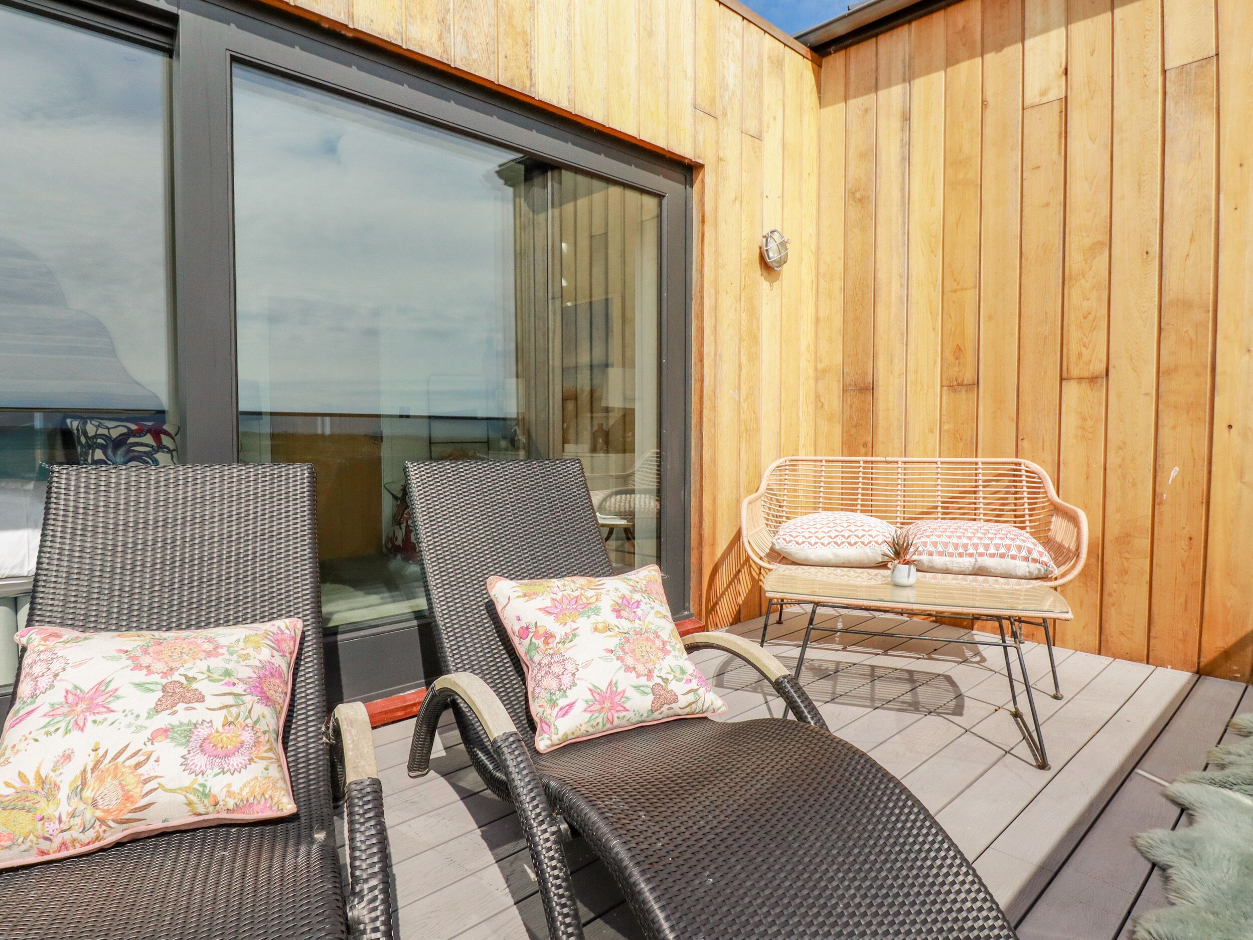 A balcony with wicker lounge chairs and a wicker table, a wooden wall, and a glass door.