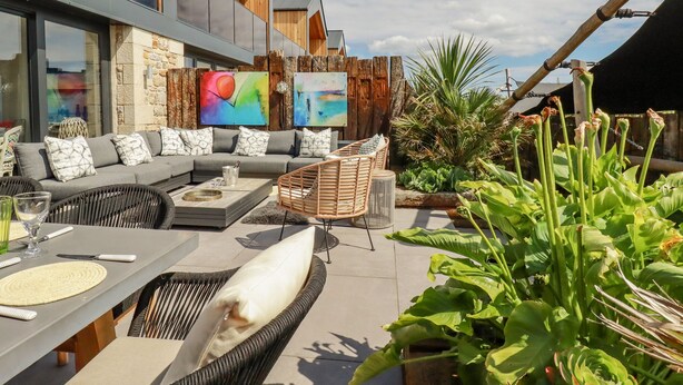 Une terrasse extérieure aménagée avec un ensemble de canapés, une table à manger et des chaises en osier. Des tableaux ornent le mur et diverses plantes sont disposées aux alentours.