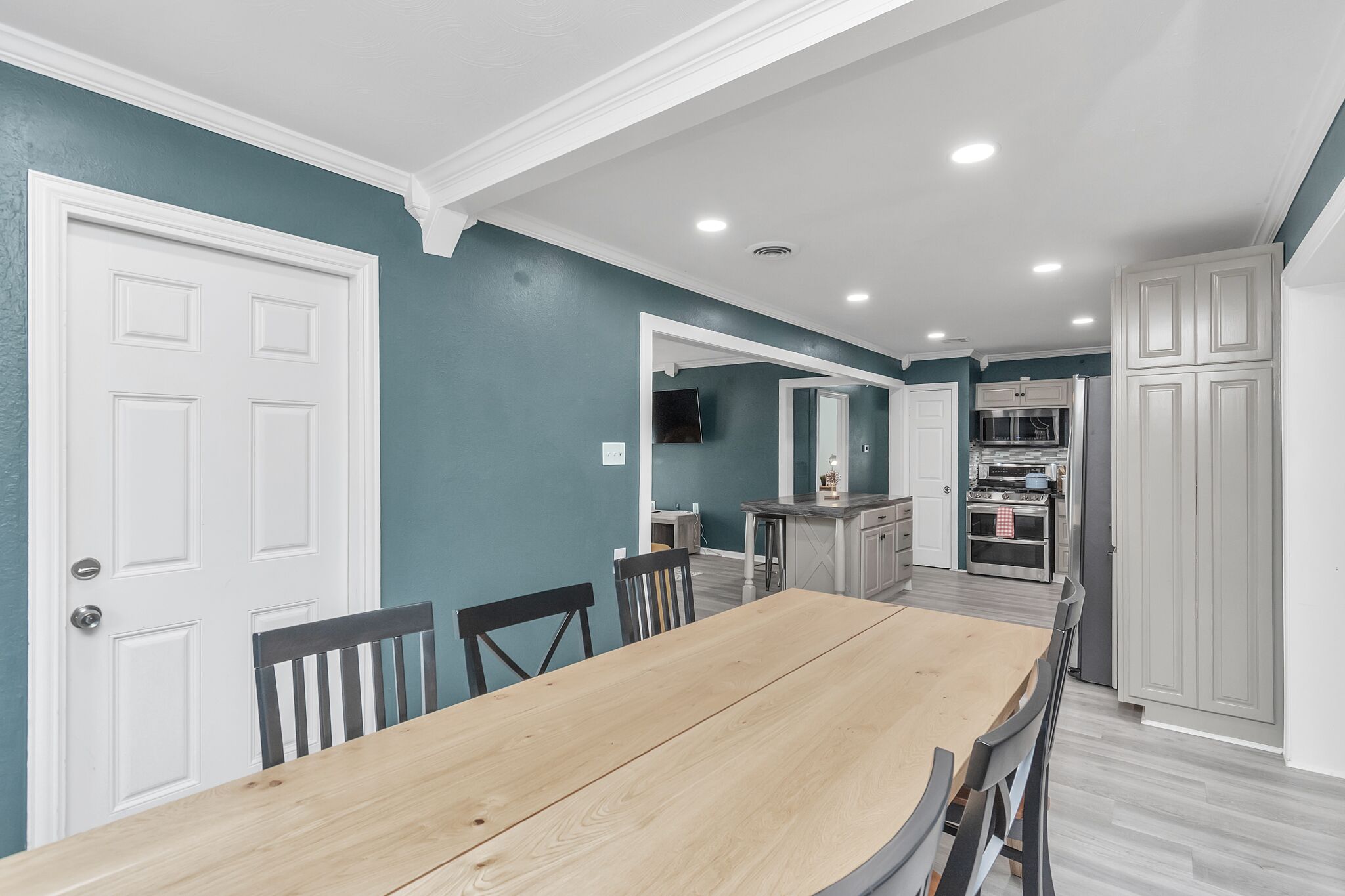 Un comedor con mesa y sillas de madera, una cocina con muebles y electrodomésticos blancos, y paredes color turquesa.