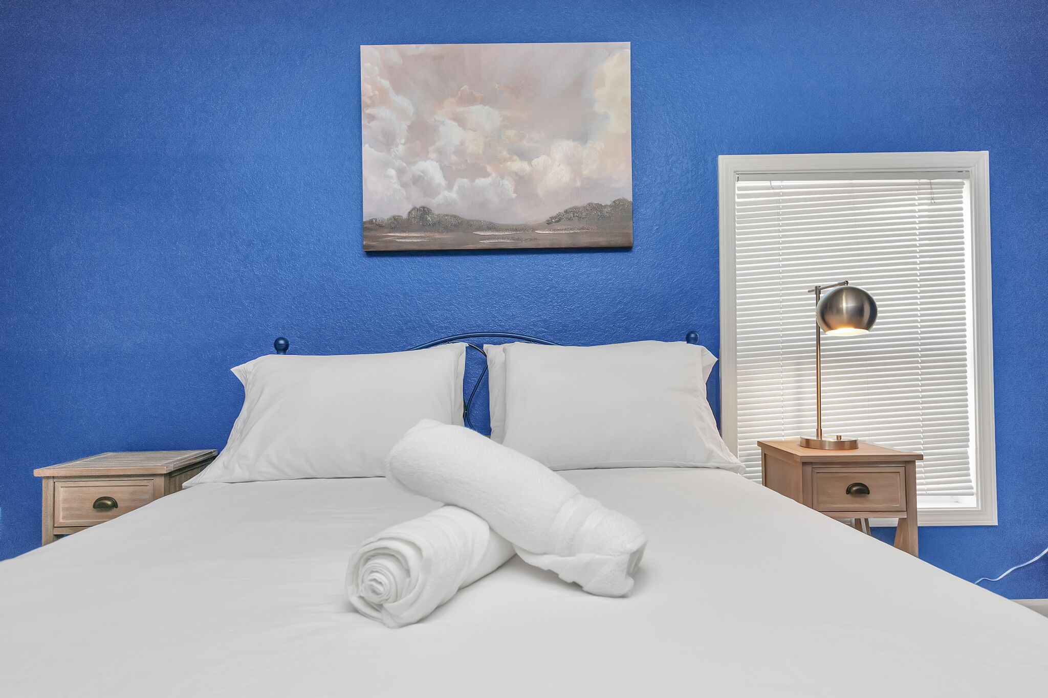 A neatly made bed with white linens and a rolled towel, a bedside table with a lamp, and a painting of a landscape on the wall.