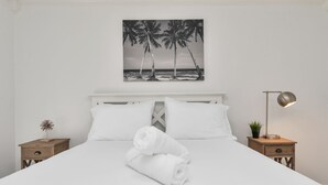 Un lit bien fait, avec du linge de lit blanc et des serviettes pliées, un tableau représentant des palmiers accroché au mur, une lampe de chevet et une petite plante posée sur la table de nuit.