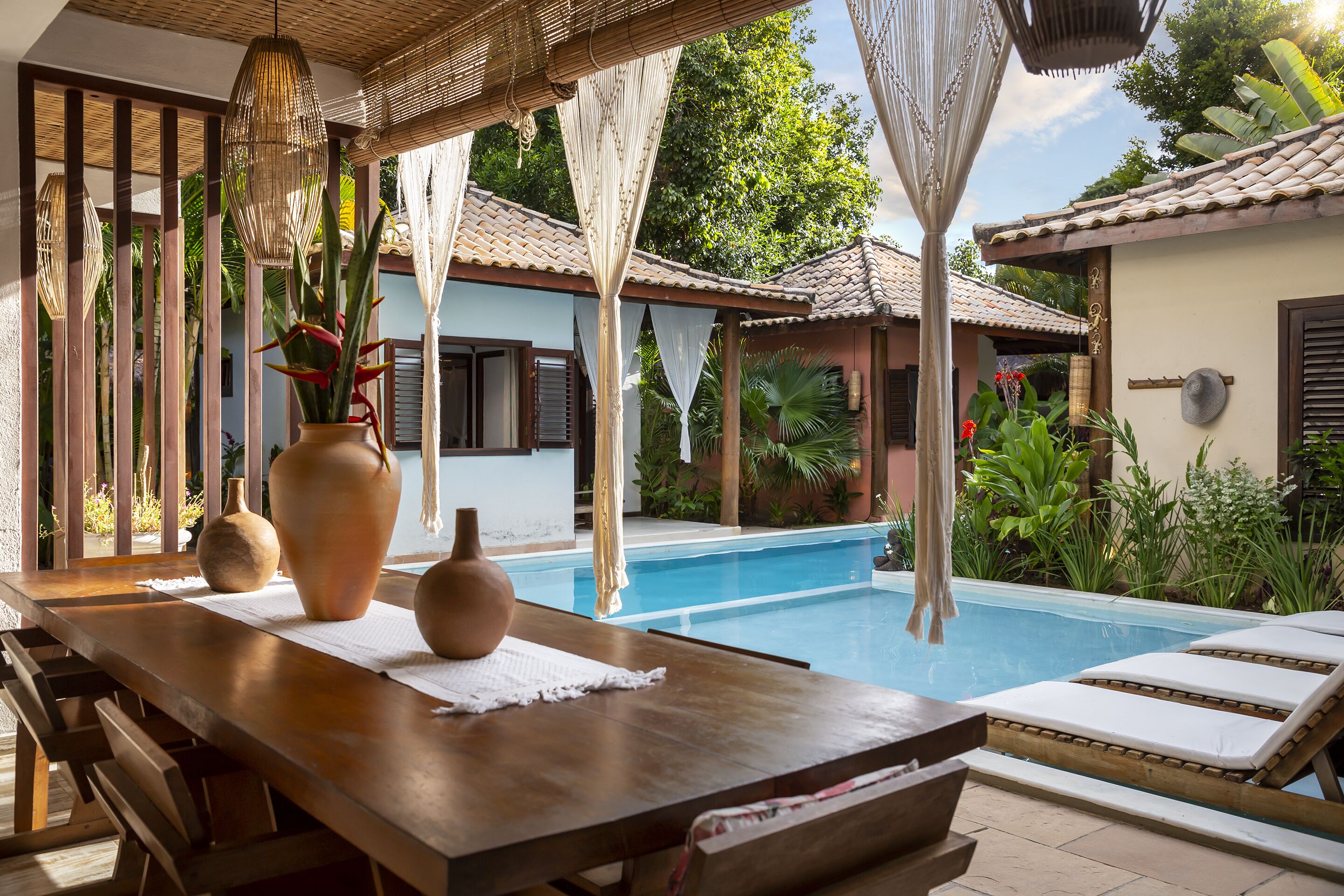 Área à beira da piscina com mesa de madeira, dois vasos grandes e uma piscina com espreguiçadeiras.