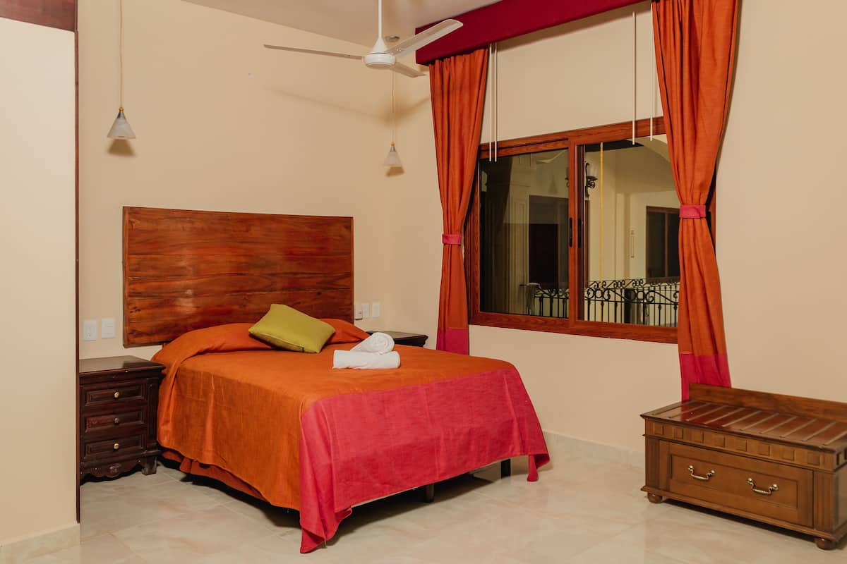 Ein Schlafzimmer mit hölzernem Kopfteil, einem Bett mit orangefarbener Bettwäsche, einem Nachttisch aus Holz, einer roten Decke, einer hölzernen Kommode und einem Fenster mit orangefarbenen Vorhängen.