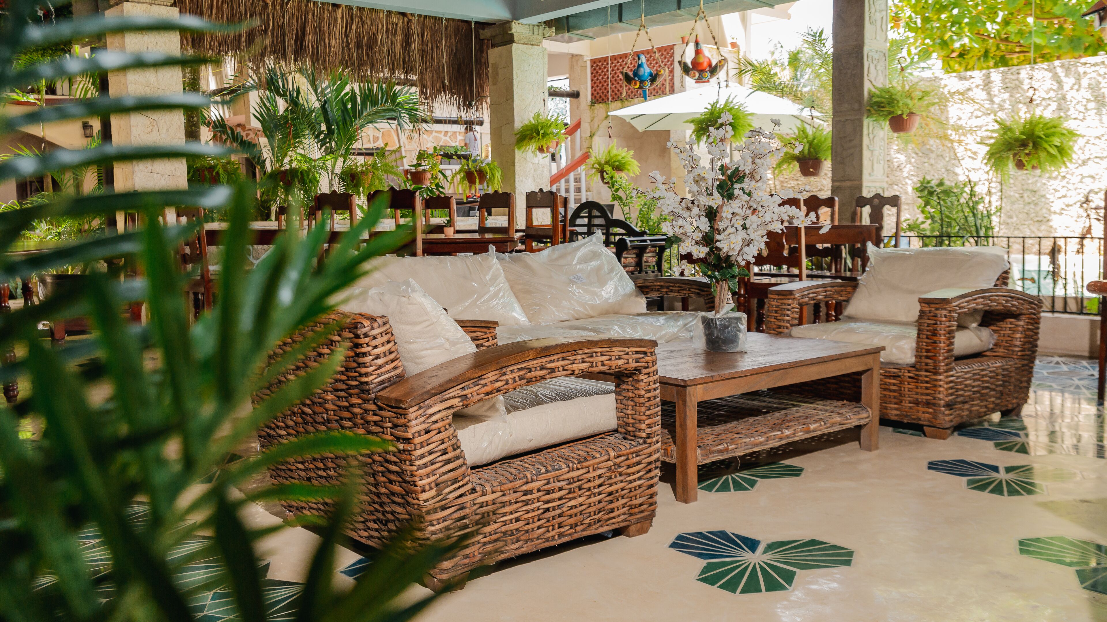 A wicker sofa set with cushions, a coffee table, and a hanging plant in a well-lit indoor space with tiled flooring.