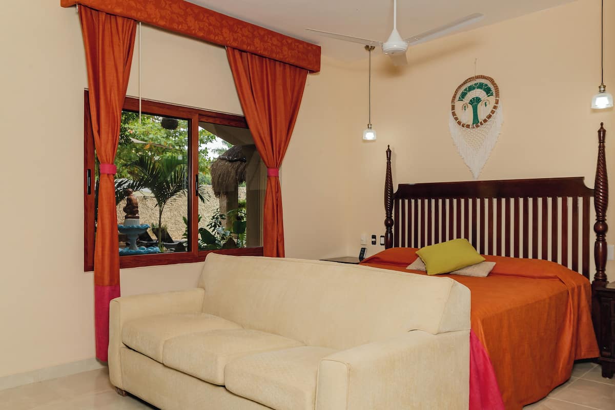 Ein Schlafzimmer mit einem beigefarbenen Sofa, einem hölzernen Bett mit orangefarbener Bettwäsche, einem Deckenventilator und einem Fenster mit orangefarbenen Vorhängen.
