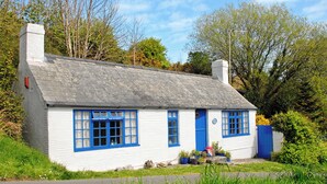 Ein charmantes weißes Cottage mit blauer Fassade und grauem Dach, umgeben von üppiger Vegetation und Bäumen.