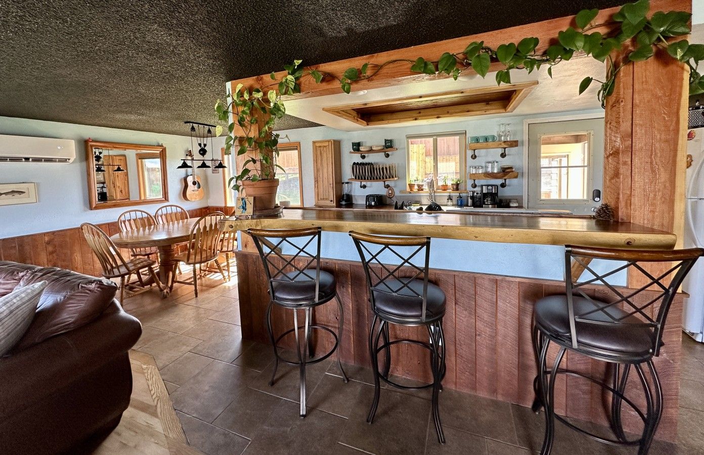 A kitchen with a wooden bar, bar stools, a dining table with chairs, and a potted plant.