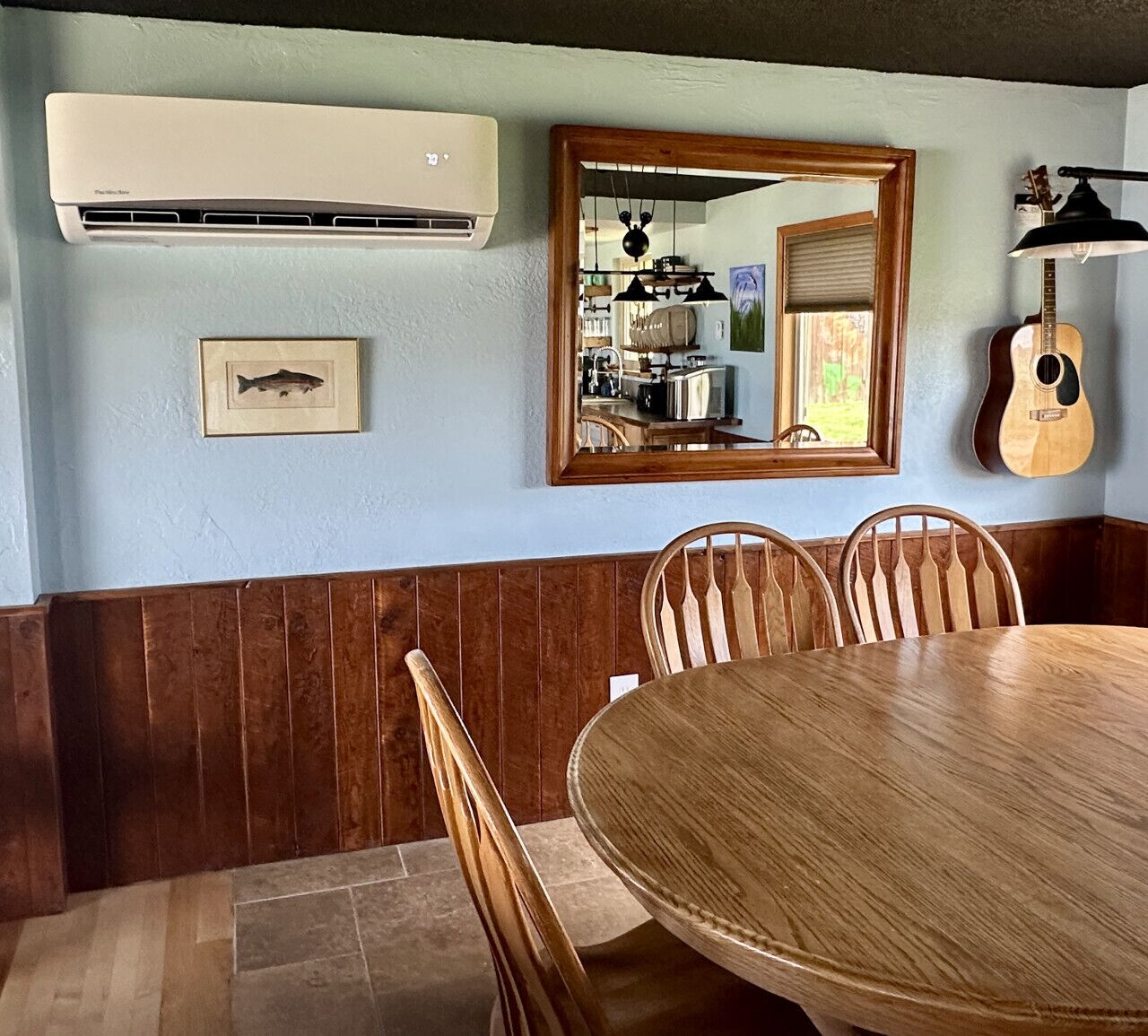 Un comedor con una mesa redonda de madera, sillas de madera, un aire acondicionado de pared, un cuadro enmarcado, un espejo y una guitarra.
