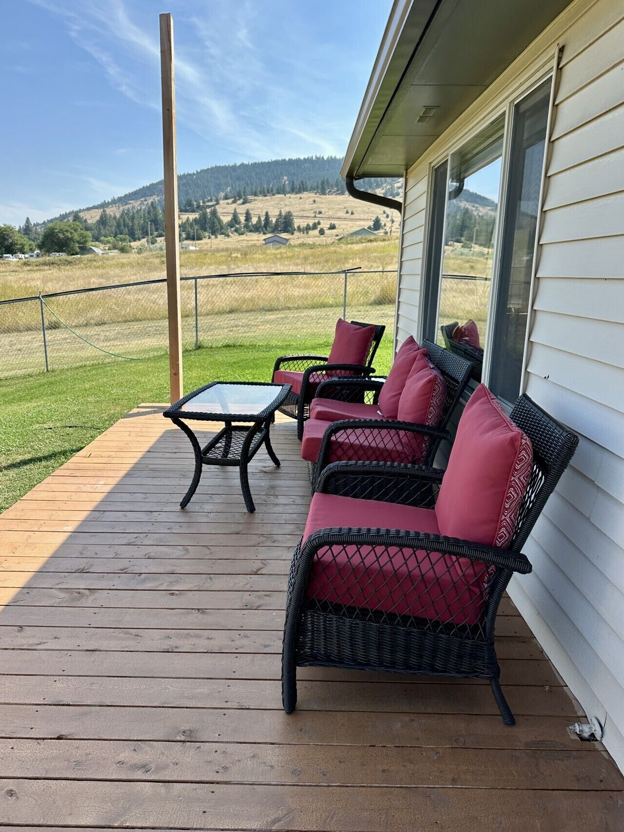 A covered outdoor seating area with wicker furniture and a glass table, overlooking a grassy field and hills.