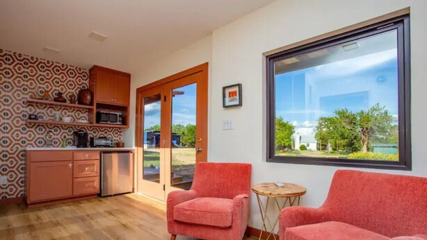 Eine moderne Küche mit Holzschränken, einer Mikrowelle und einem Kühlschrank. Zwei rote Stühle und ein kleiner runder Tisch bieten Platz. Ein großes Fenster gibt den Blick auf Grünflächen frei.