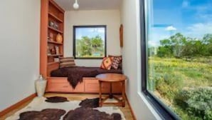 Ein gemütliches Zimmer mit großem Fenster, einem Bett, einem kleinen Tisch und einem Bücherregal.