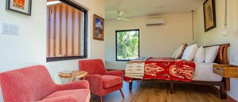 A bedroom with a bed, two red armchairs, a small round table, and a window with blinds.