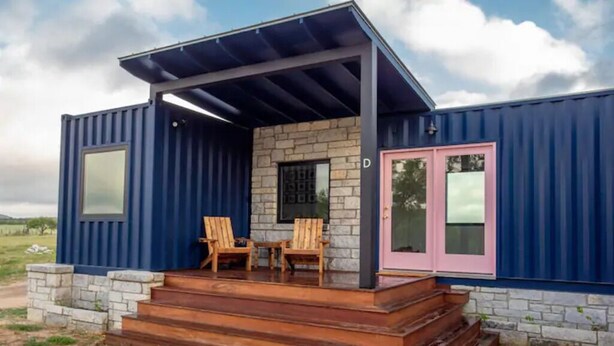 Een moderne containerwoning met een stenen gevel, een overdekte veranda en een roze deur.