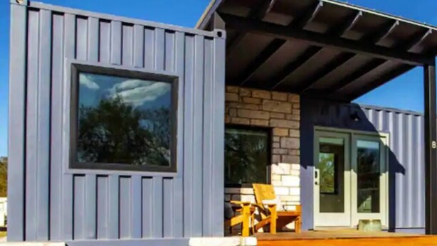 A modern building with a large window, a stone facade, and a covered porch with outdoor seating. - Tranquility at Odonata Escape --- Chic modern container home in a country setting (Fredericksburg)
