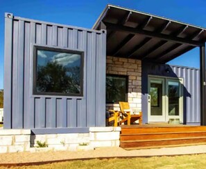 A modern building with a large window, a stone facade, and a covered porch with outdoor seating. - Tranquility at Odonata Escape --- Chic modern container home in a country setting (Fredericksburg)