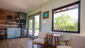 A modern kitchen with wooden cabinetry, a microwave, and a refrigerator. There are two chairs and a small round table. A large window provides a view of trees outside. - Tranquility at Odonata Escape --- Chic modern container home in a country setting (Fredericksburg)