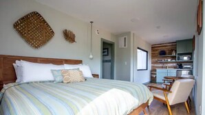 A bedroom with a large bed, a small desk, and a kitchenette. - Tranquility at Odonata Escape --- Chic modern container home in a country setting (Fredericksburg)