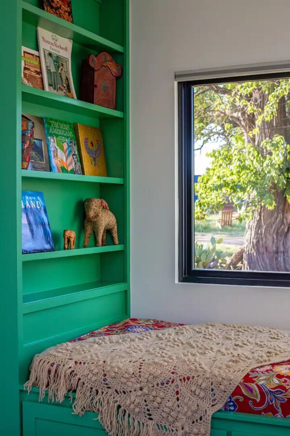 A room with a green bookshelf, a window with a view of trees, and a bed with a patterned blanket. - Happy Boho at Odonata Escape -- Luxurious Shipping Container stay in a rural setting (Fredericksburg)