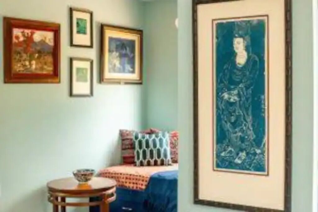 A room with a bed, a round table with a bowl, and framed artwork on the wall.