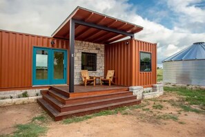 Een laag, klein gebouw met een veranda, een blauwe deur en een kleine tafel met stoelen.