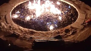 Ein runder Feuerkorb im Freien mit Flammen, umgeben von Holzstühlen.