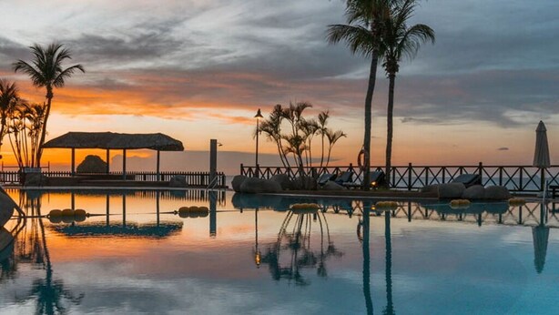 Una piscina infinita tranquila al atardecer con una terraza de madera, palmeras y un pabellón.