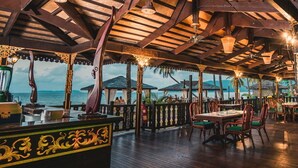 Un restaurante al aire libre con mesas y sillas de madera, vista al océano y una zona cubierta con luces colgantes.
