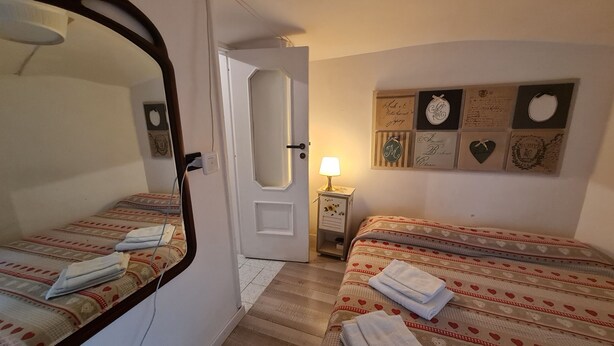 Une chambre avec deux lits, une table de chevet avec une lampe, un mur orné de tableaux ou d’objets décoratifs, et une porte.