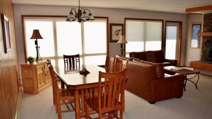 A spacious living room with a wooden dining table, two chairs, a brown leather sofa, and a television. - 2 Bedroom + Sleeping loft condo near mountain village w/shuttle route (Gallatin Gateway)