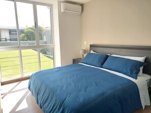 A bedroom with a large bed, a nightstand, a lamp, and a view of a green lawn through a large window.