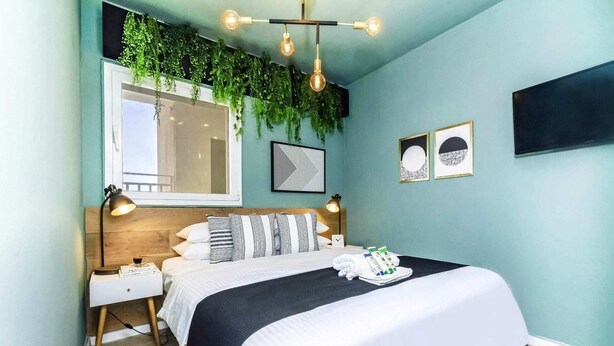 A bedroom with a bed, bedside tables, a wall-mounted light fixture, a window, and a wall with framed artwork. - Dorot 13 - Studio Apartment, Sleeps 2 (Jerusalem)